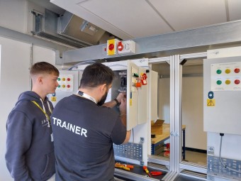 A training instructor shows electrical equipment to a younger student while working on a machine in a workshop.