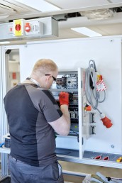 A man in a black shirt and red gloves is focused on working on a control panel.