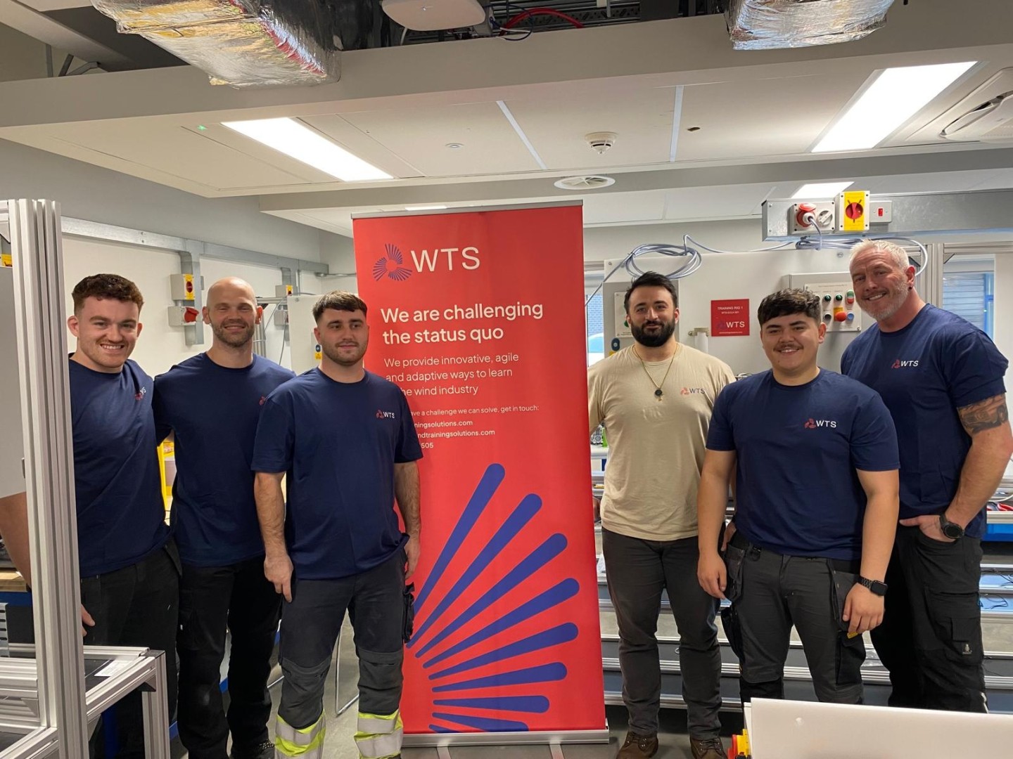 An image of 5 men in blue t-shirts and 1 man in a sand coloured t-shirt standing between a red roller banner that advertises WTS services and how they are changing the status quo. 