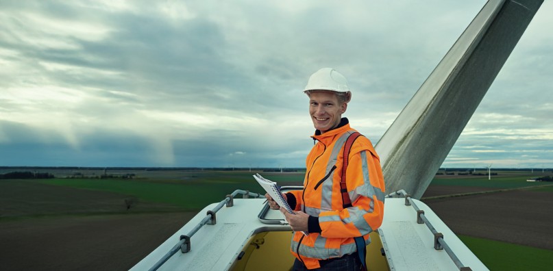 A wind turbine technician in an orange vest and hard hat holds a notebook atop a wind turbine at a wind farm.